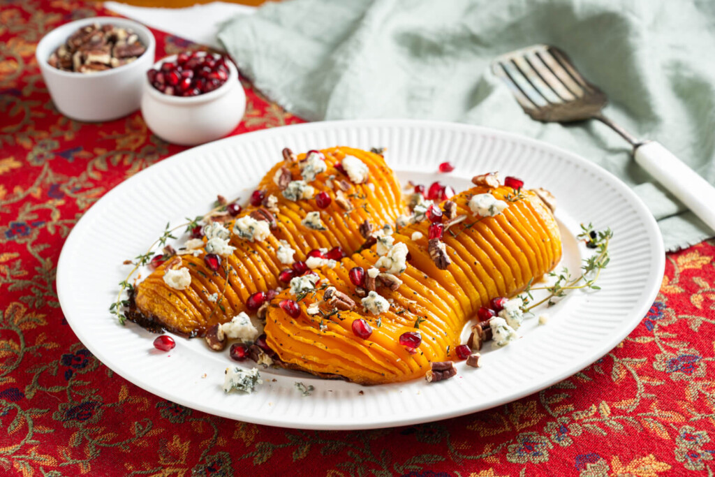 Hasselback roasted butternut squash on a white plate garnished with pomegranate arils, toasted pecans, and blue cheese.