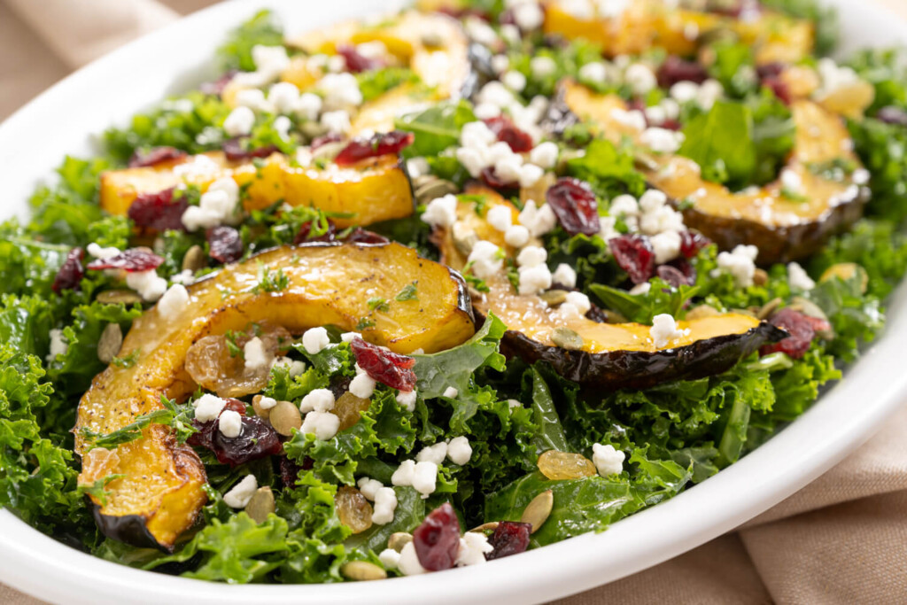 Serving dish of roasted acorn squash and kale salad, garnished with goat cheese, dried cranberries, and pumpkin seeds.