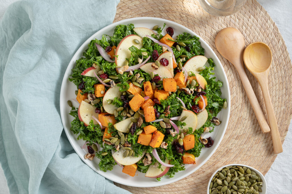 Above view of warm kale, butternut squash, and apple salad garnished with red onions, cranberries, golden raisins, and pumpkin seeds.