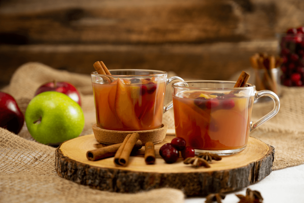 The images shows two clear mugs filled with mulled apple cider on a cross-section of leg. The mugs and log are garnished with cinnamon sticks and star anise. In the left background are red and green apples.