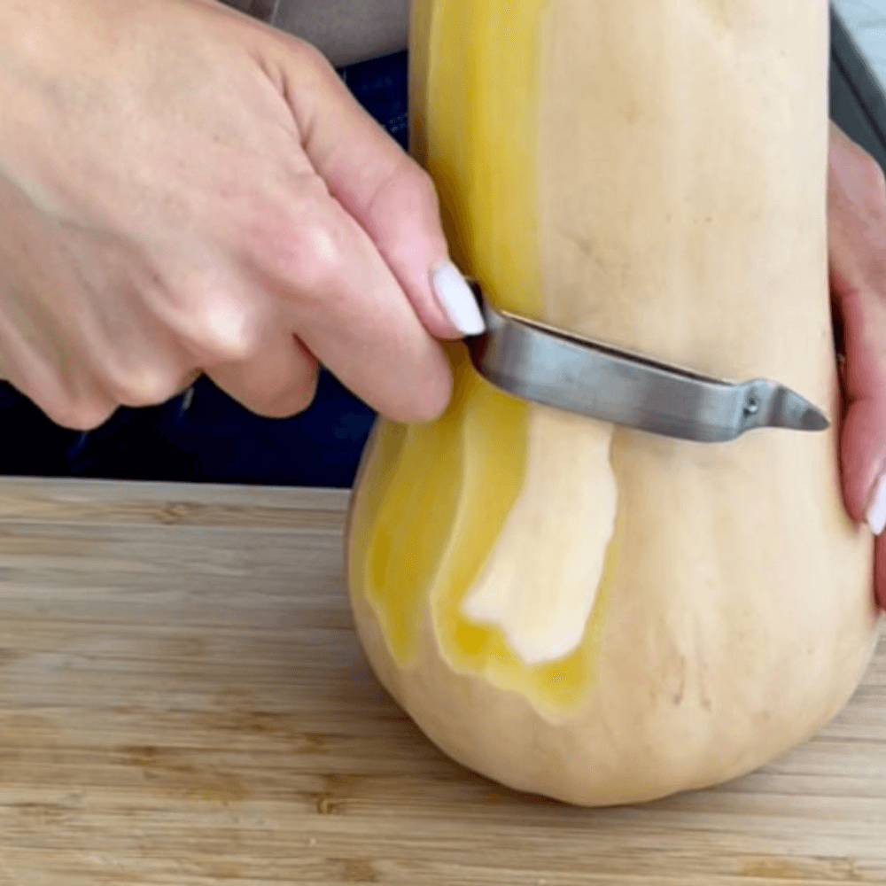 Hand peeling butternut squash on wooden cutting board with vegetable peeler.