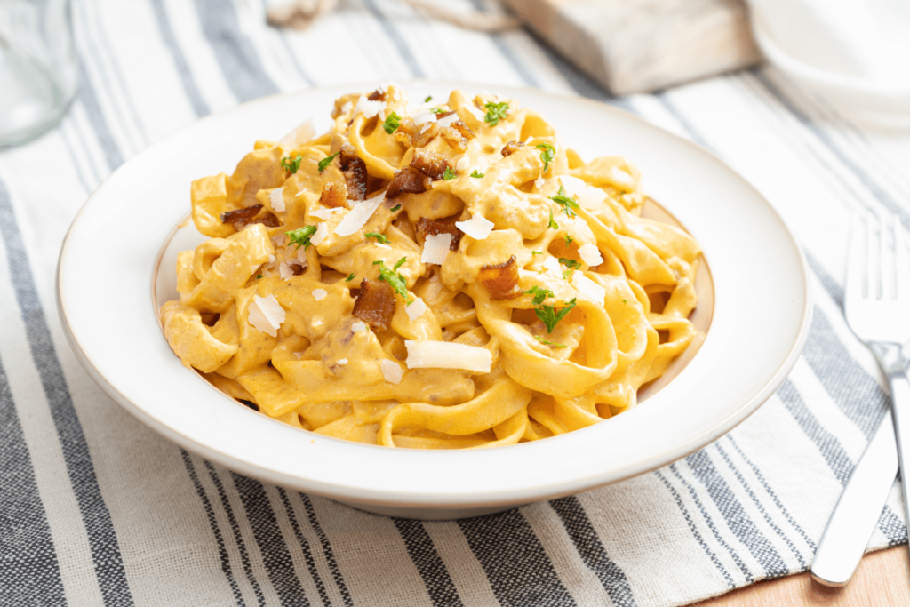 White dish of creamy pumpkin chick tagliatelle garnished with bacon bits and Parmesan cheese on a striped placemat.