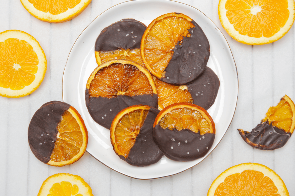 Overhead shot of shite plate with chocolate covered dried oranges surrounded by sliced fresh oranges.