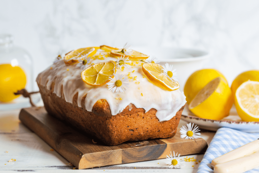 Lemon loaf cake on wooden cutting board with white icing, dried lemons, and small daisy flowers.