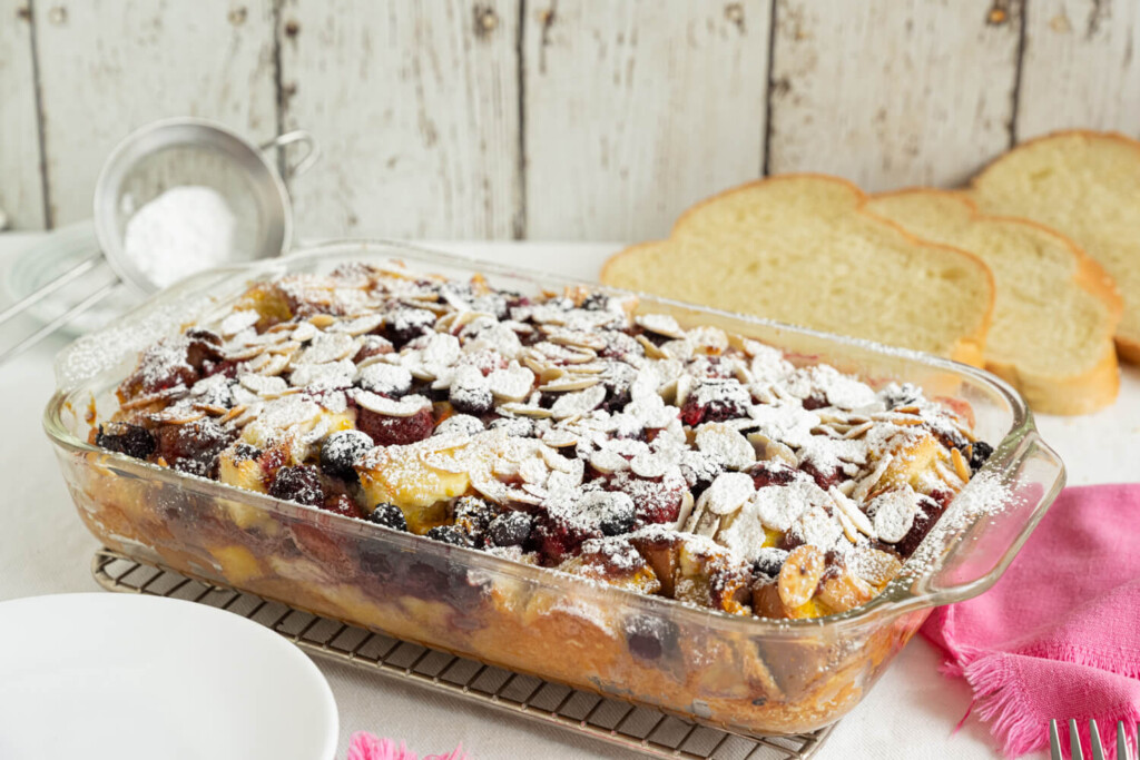 Glass dish with mixed berry strata dusted with icing sugar. In the background are three slices of challah bread.