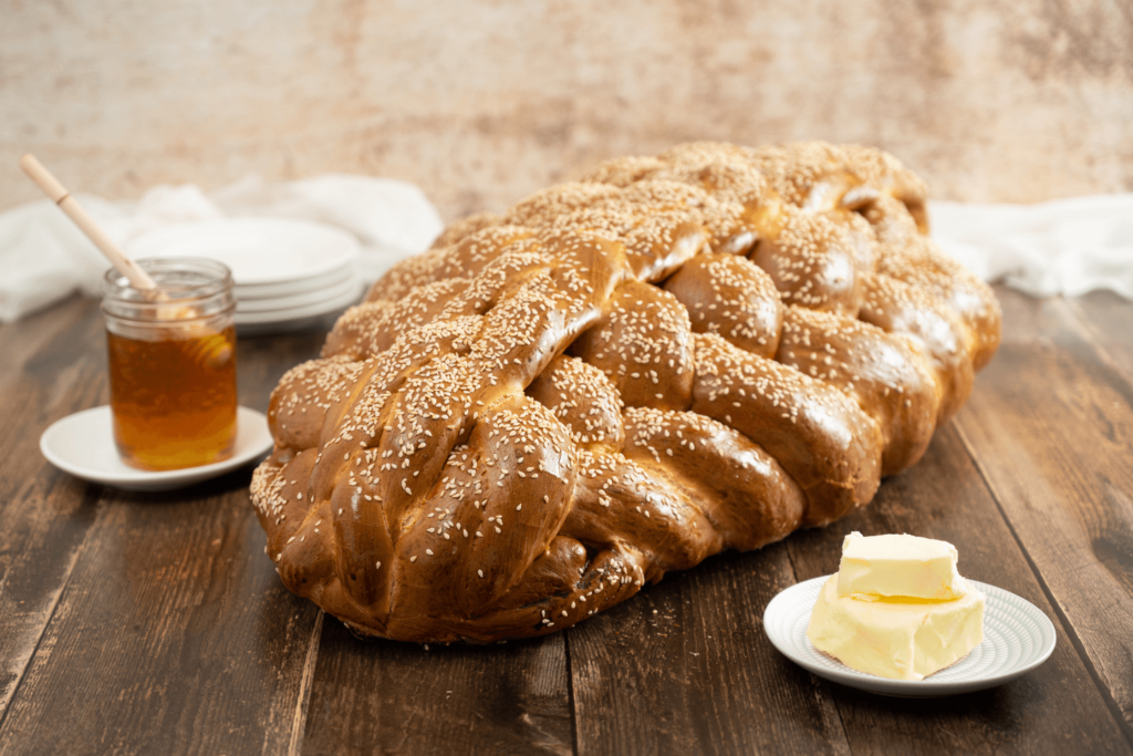 Full loaf of 8-braid challah bread on dark wood table between jar of honey and block of butter.