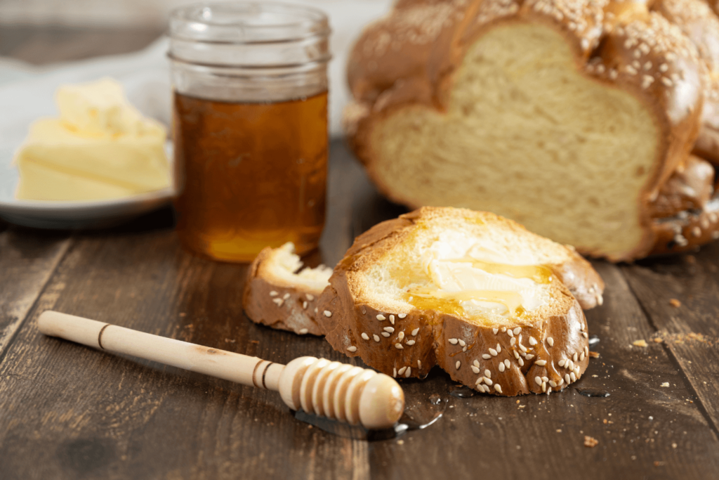 Sliced loaf of challah bread with slice drizzled with honey on dark wood table.