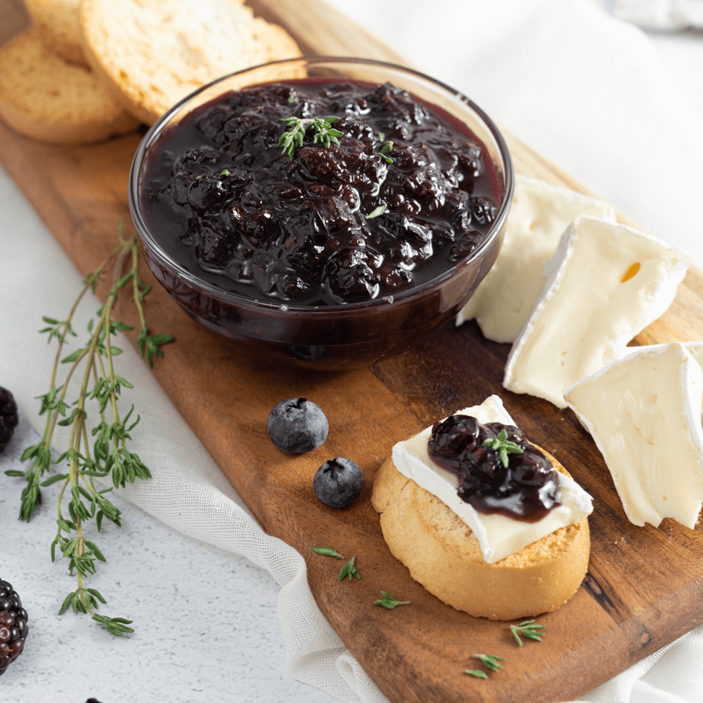 Holiday Appetizer Recipe: a wooden serving plank with a small bowl of dark berry compote behind a slice of baguette with a wedge of Brie and a spoonful of compote.