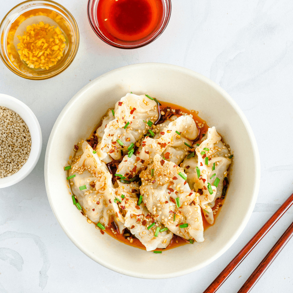 Overhead image of a white bowl filled with dumplings garnished with fresh chilis, green onions, and sesame seeds.