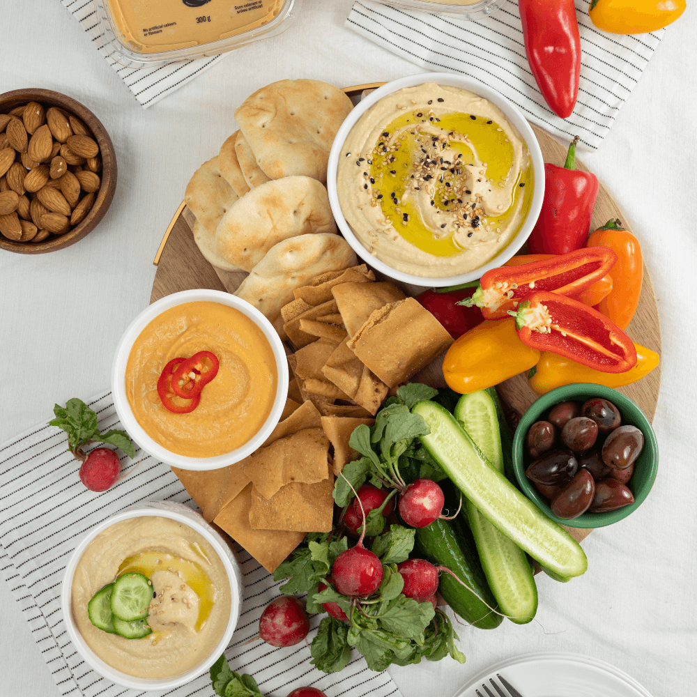 Overhead image of serving platter filled with fresh cut veggies and chips with three bowls containing different flavours of Farm Boy Hummus.