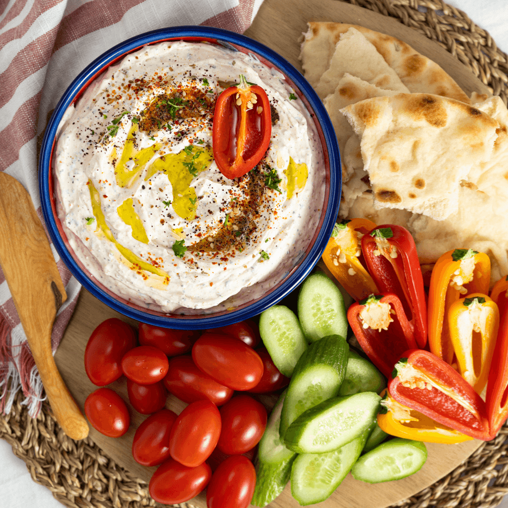 Holiday Appetizers Recipe: Overhead image of a bowl of spiced labneh dip finished with olive oil ad surrounded by fresh cut veggies and torn pieces of naan bread.