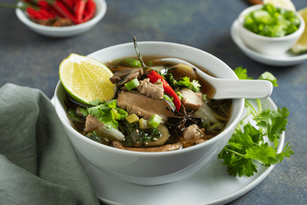 Bowl of warm chicken soup with Asian greens garnished with Thai chili peppers, green onions, and lime.