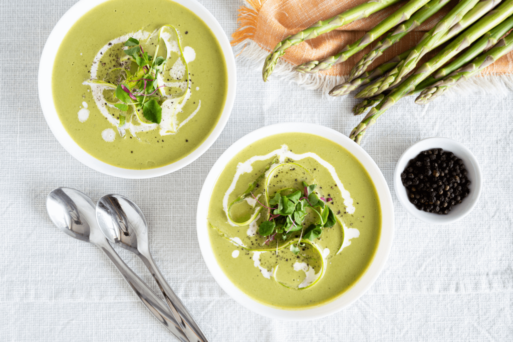 holiday leftovers: The image is show from above and shows two bowls of creamy asparagus soup, garnished with swirls of whipped cream, asparagus ribbons, cracked black pepper, and microgreens. In the upper right corner are stalks of fresh asparagus on a peach-coloured napkin.