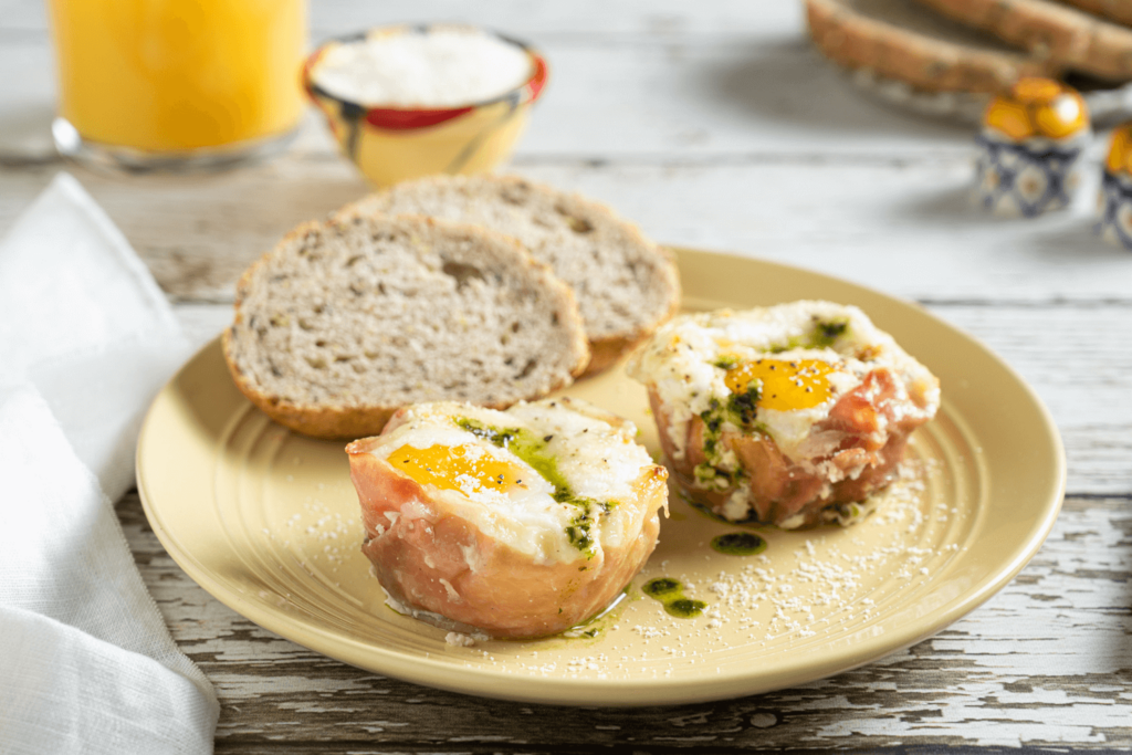 Easy Breakfast Idea: The image shows a pale yellow plate with two slices of bread and two prosciutto egg cups with cheese and pesto.