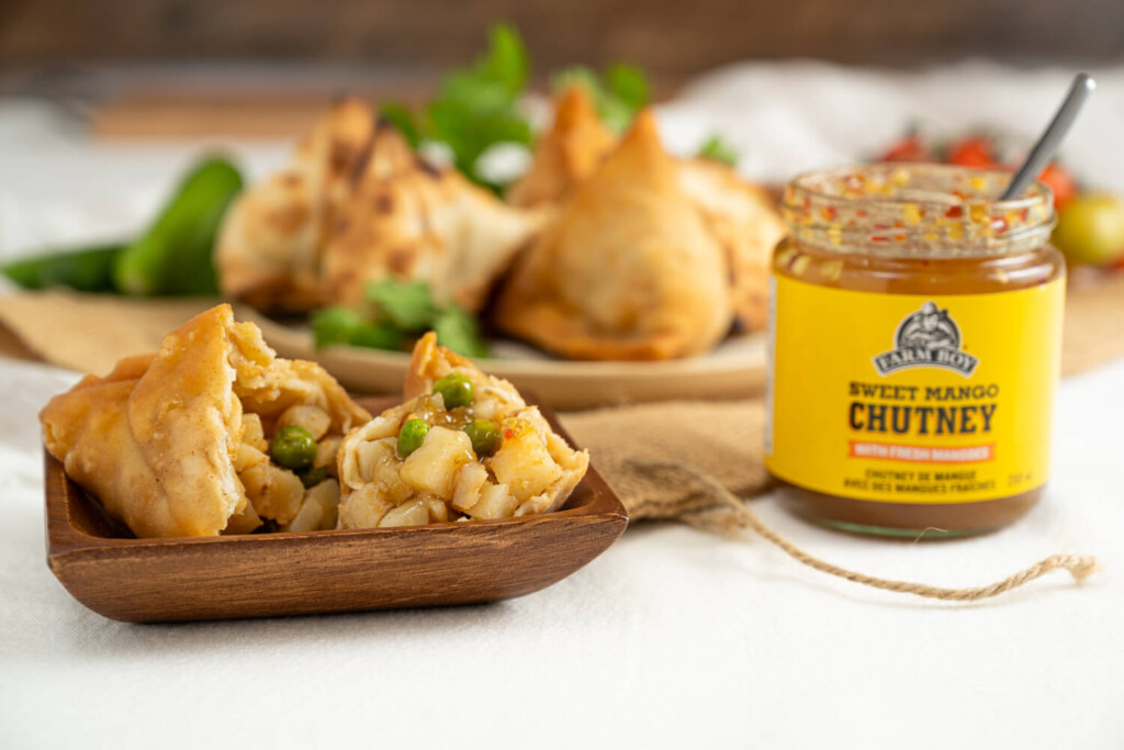 The image shows a small square wooden plate in the foreground with a samosa cut in half. Visible inside the samosa are chunks of potatoes and peas. Beside the square plate is an opened jar of Farm Boy Sweet Mango Chutney. There is a spoon in the jar. In the background, out of focus are more samosas on a wooden serving plate.