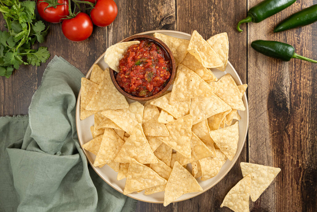 The image is taken from above and shows a large white plate with tortilla chips. At the 11 o'clock position of the plate is a small bowl with fresh tomato salsa. In the upper right corn of the image are three jalapeno peppers. In the upper left corner of the image is fresh cilantro and three tomatoes on the vine. In the lower left corner of the image is a sage green piece of cloth.