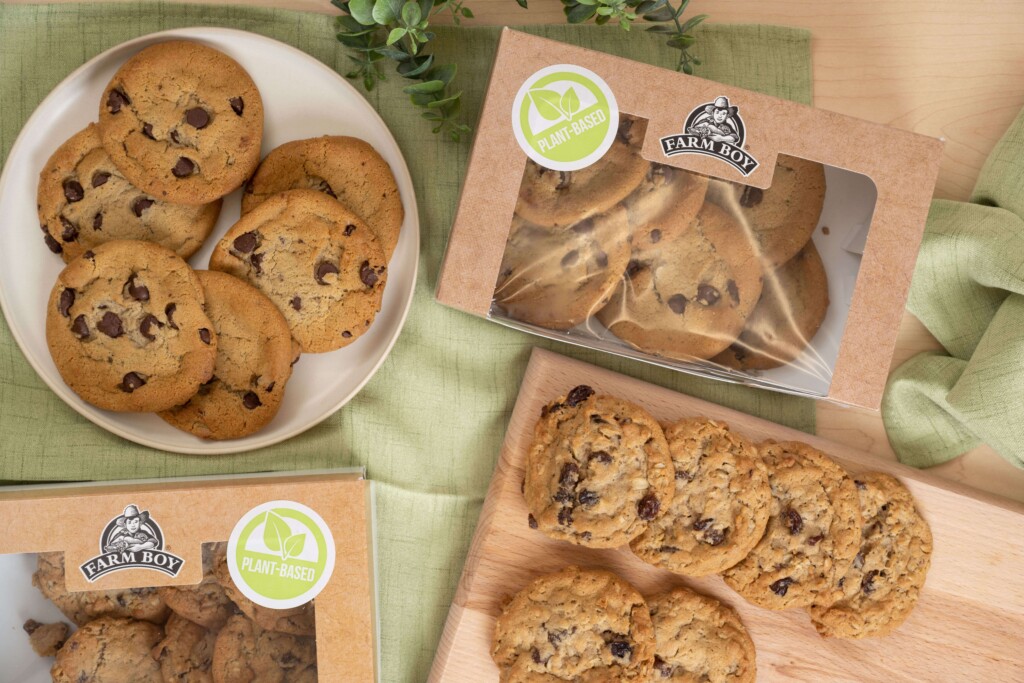 The image is shot from above and shows a light wood table with a pale green tablecloth. In the upper right corn is a box of Farm Boy Plant Based Chocolate Chip Cookies. Below the box is a wooden cutting board with two rows of oatmeal raisin cookies. To the left of the box is a white plate with chocolate chip cookies. Bwlo the plate is the corner of another box, with the Plant Based sticker visible.