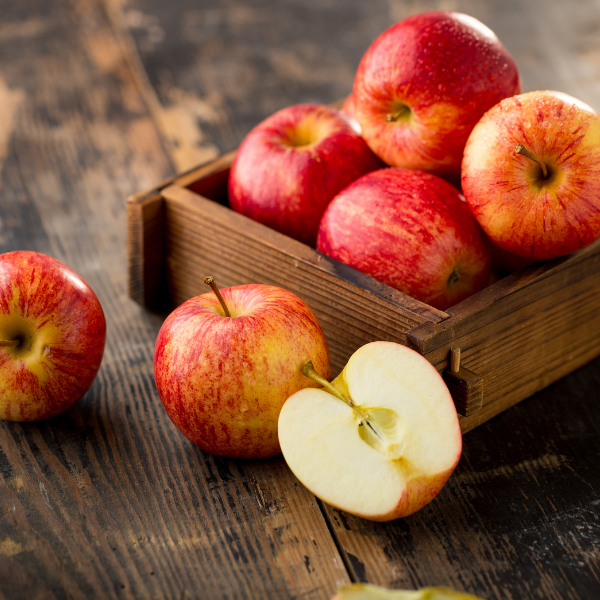 The image shows a wooden table with a small wooden crate on it. Visible in the crate are four reddish-yellow apples. IN front of the crate is a whole apple and an apple sliced in half. To the left side of the image beside the crate is another whole apple.