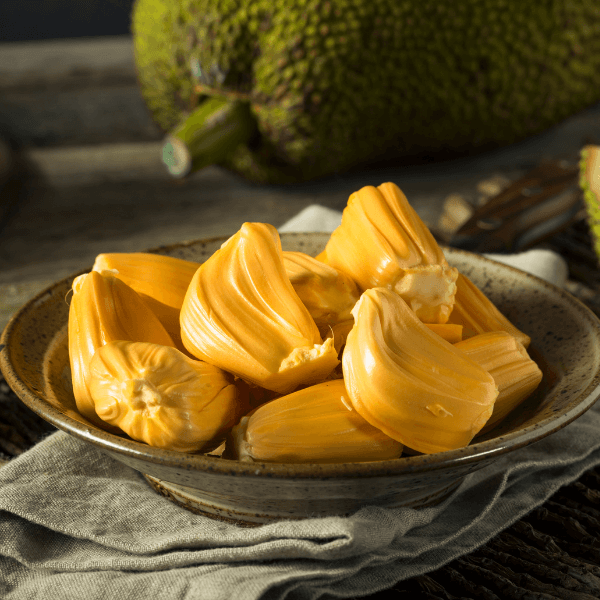 The image shows a grey tablecloth folded over and a speckled stoneware bowl on top. In the bowl are segments of fresh jackfruit. In the background out of focus, is a whole jackfruit lying on its side.