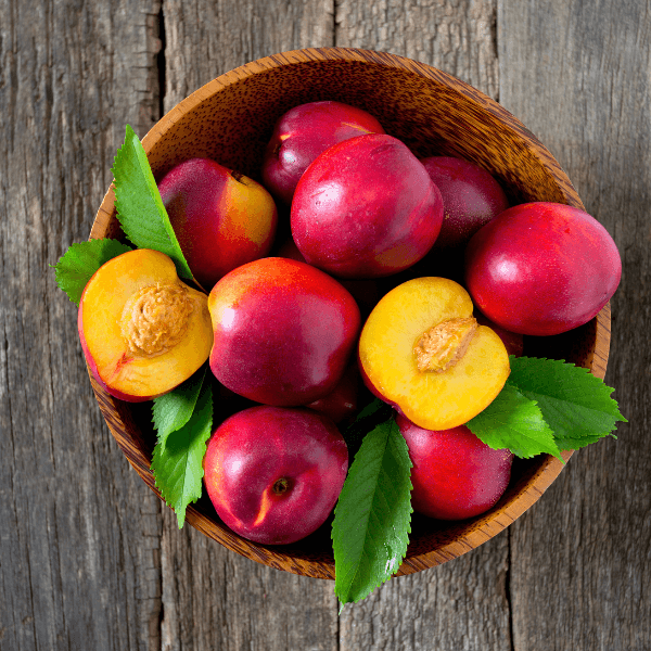 The image is shot from above and shows a grey wood table with a wooden bowl. In the bowl are fresh nectarines. Two of the nectarines are cut in half, with their pits visible. The nectarines are bright and vibrant with shiny skins.