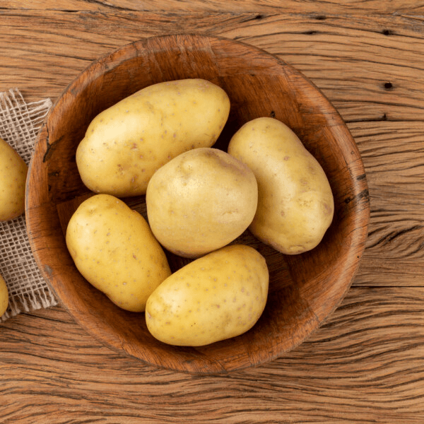 The images shows a wooden table with a medium-sized wooden bowl on it. In the bowl are five medium-sized Yukon Gold potatoes.