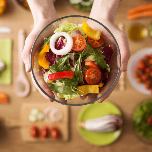 Overhead shot of hands holding glass bowl of salad above table laid with cutting boards, chopped vegetables, and utensils.