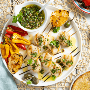 Grilled scallops with salsa verde on white plate with red and yellow peppers and grilled lemons. Easy seafood appetizer recipe.