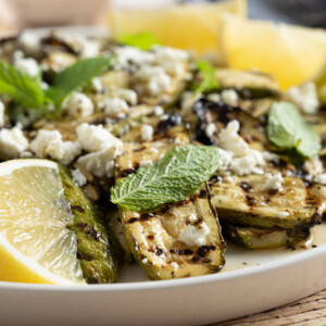 A close-up of a plate of grilled zucchini slices topped with crumbled feta cheese, mint leaves, and lemon slices.