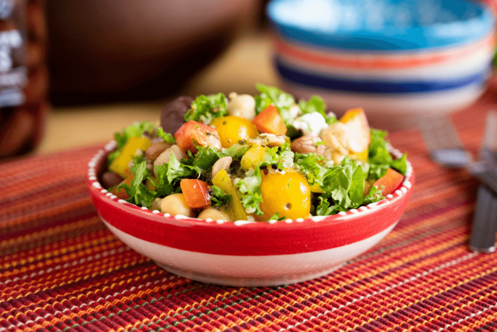 The image shows a red woven placement. Out of focus in the background are stacked bowls in blue, red, and white. In the foreground is a red and white bowl. In the bowl is a salad of chopped kale, yellow bell peppers, tomatoes, olives, and quinoa.