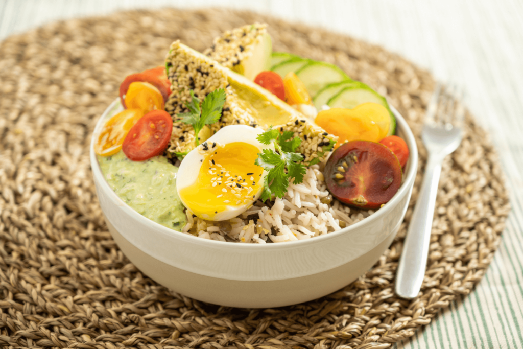 Cold Dinner Idea: Eda-Yummy Grain Bowl. The image shows a round wicker placemat with a white bowl in it. Beside the bowl is a fork. In the bowl is rice. On top of the rice is sliced cucumber coins, halved cherry tomatoes, a soft-boiled egg, and wedges of avocados. The avocados and egg have Farm Boy's Everything Bagel Seasoning Blend.