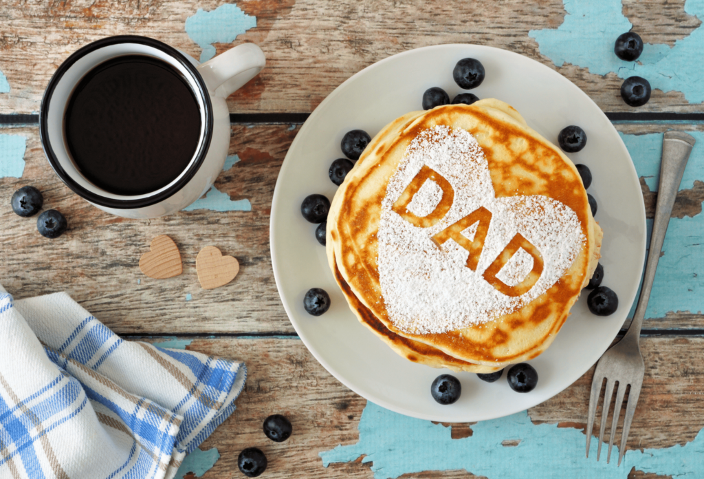 Father's Day breakfast. plate of pancakes with DAD in icing sugar, surrounded by blueberries. a mug of coffee and a blue plaid dish towel are also on the table