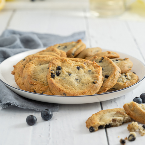 The image shows a white b=plate with a black rim lying partially on a slate blue cloth and on a white-washed wood table. On the plate, arranged in a fanned circle are lemon blueberry cookies. In the foreground lower right corner is a lemon blueberry cookie in two halves, with some crumbs. There are two fresh blueberries behind this cookie. To the lower centre left of the plate are two more fresh blueberries.