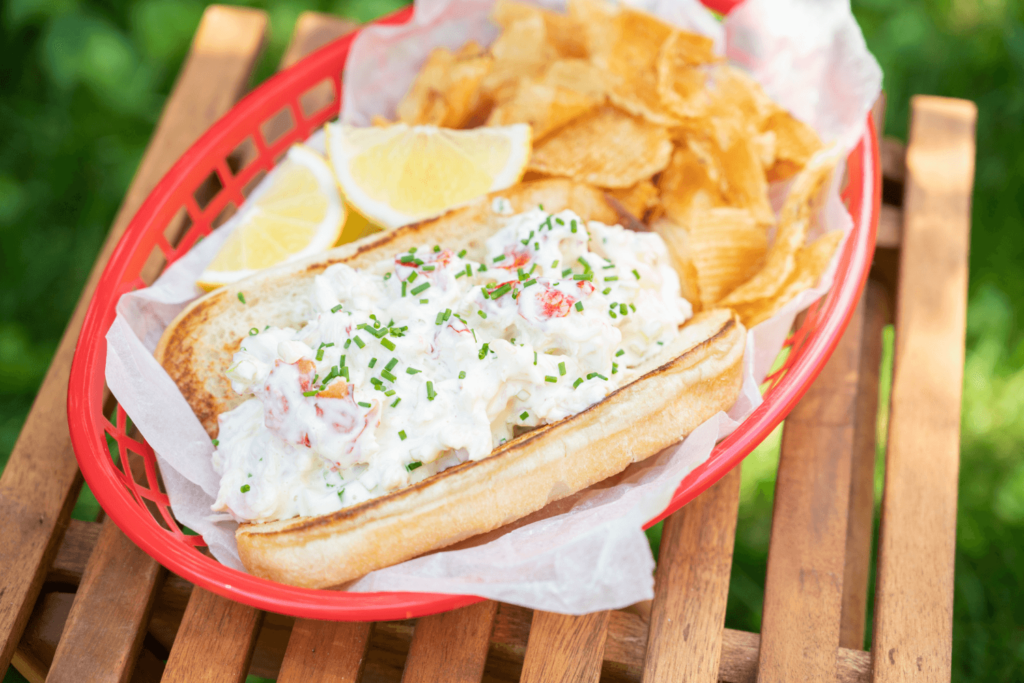 Cold Dinner Idea: Lobster Roll. The image shows a slatted wooden table with a red plastic fry basket on it. In the fry basket is a hot dog bun loaded with lobster meat, mayonnaise, and a sprinkle of fresh chives. Atop the roll are two wedges of lemon and beside it are Farm Boy Crinkle Cut Potato Chips