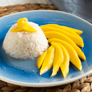 The image shows a blue plate on a round wicker placemat. On the plate is a mound of rice with a twist of fresh mango on top. Thin slices of mango are fanned out next to the mound of rice. There is a grey-blue scarf behind the plate.
