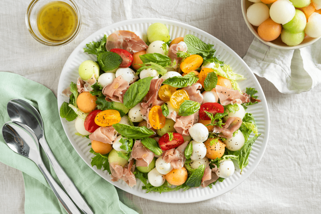 The image is taken from above. The image shows a white tablecloth with a large white plate in the centre. On the plate is a salad of romainc lettuce, halved red and yellow cherry tomatoes, balls of bocconcini cheese, sliced prosciutto, and balls of cantaloupe, honeydew, and canary melons. The salad is garnished with mint leaves and pepper. In the upper right corner of the image is a bowl filled with the balls of three melons. In the bottom left of the image is a mint green napkin with two large serving spoons. In the upper left of the image is a small clear glass bowl with an olive-oil based dressing.