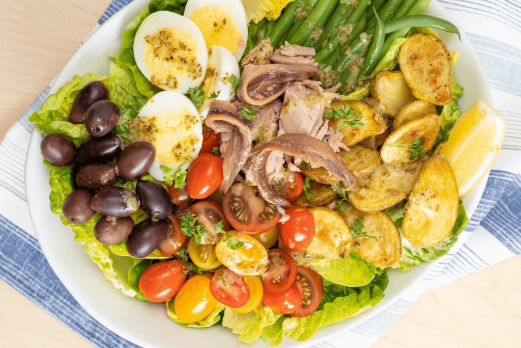 Cold Dinner Idea: Nicoise Salad. The image shows a white plate on top of a blue and white striped dishcloth. On the plate, beginning at the top and going clockwise are green beans, fried potatoes, a wedge of lemon, halved red and yellow cherry tomatoes, black Kalamata olives, and halved hard boiled eggs. In the centre is tuna and anchovies.