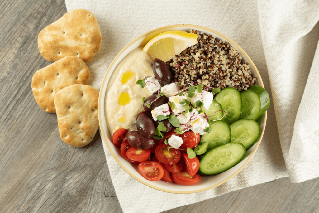 The images shows a grey wood table with a cream linen tablecloth partially draped across it. On the tablecloth is a cream coloured bowl. Clockwise from the top of the bowl is a wedge of lemon, mixed quinoa, sliced cumber, halved red cherry tomatoes, black Kalamata olives, hummus with three dots of olive oil, and cubed feta cheese with microgreens in the centtre. To the left of the bowl are three Farm Boy Naan Bites.