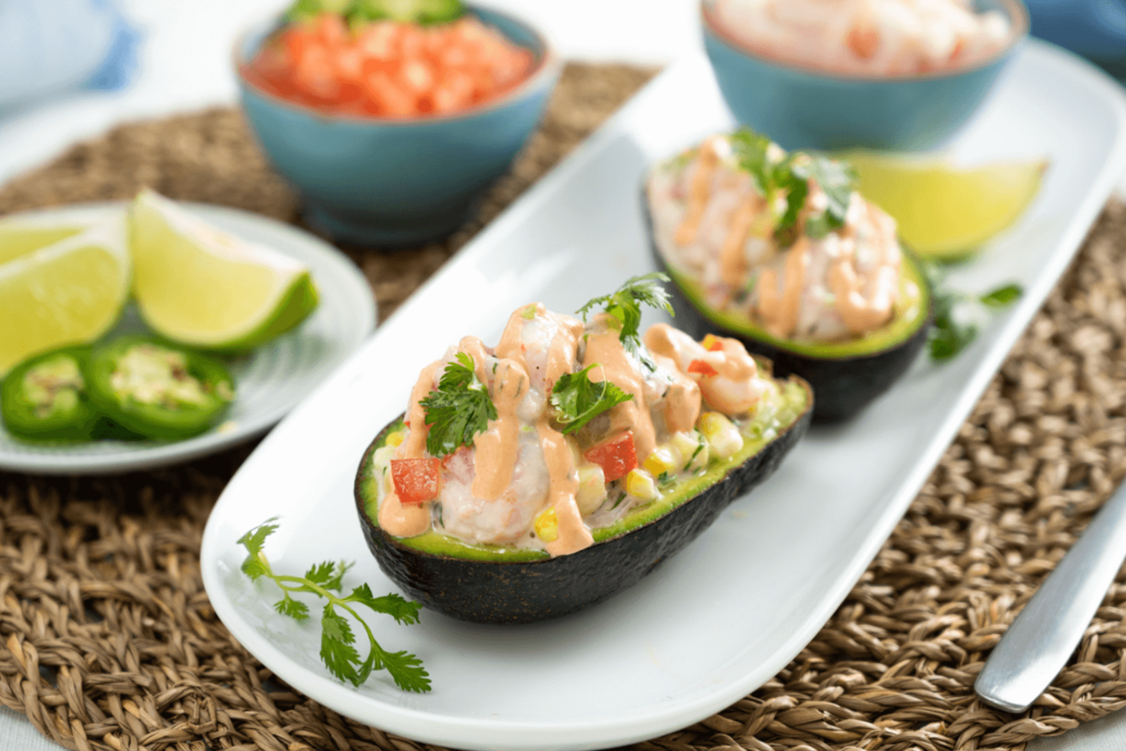 Cold Dinner Idea: Shrimp Salad Stuffed Avocados. The image shows a round wicker placemat. On top of the placemat is an oblong white dish. On the dish are two avocado halves piled with shrimp salad, drizzled with Farm Boy Chipotle Mayonnaise and garnished with fresh cilantro. Behind the avocado halves is a wedge of lime and a small blue bowl, out of focus. To the right of the oblong dish is a small plate, out of focus, with wedges of lime and sliced fresh jalapeno pepper. Behind the plate and out of focus is a small blue bowl filled with chopped tomatoes.