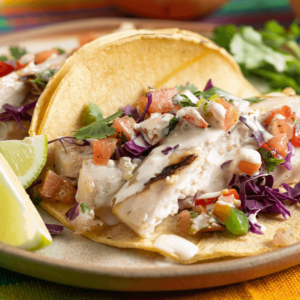 Grilled fish tacos made with mahi mahi, topped with fresh salsa, cabbage, and a creamy sauce, served with lime wedges on a colorful placemat.