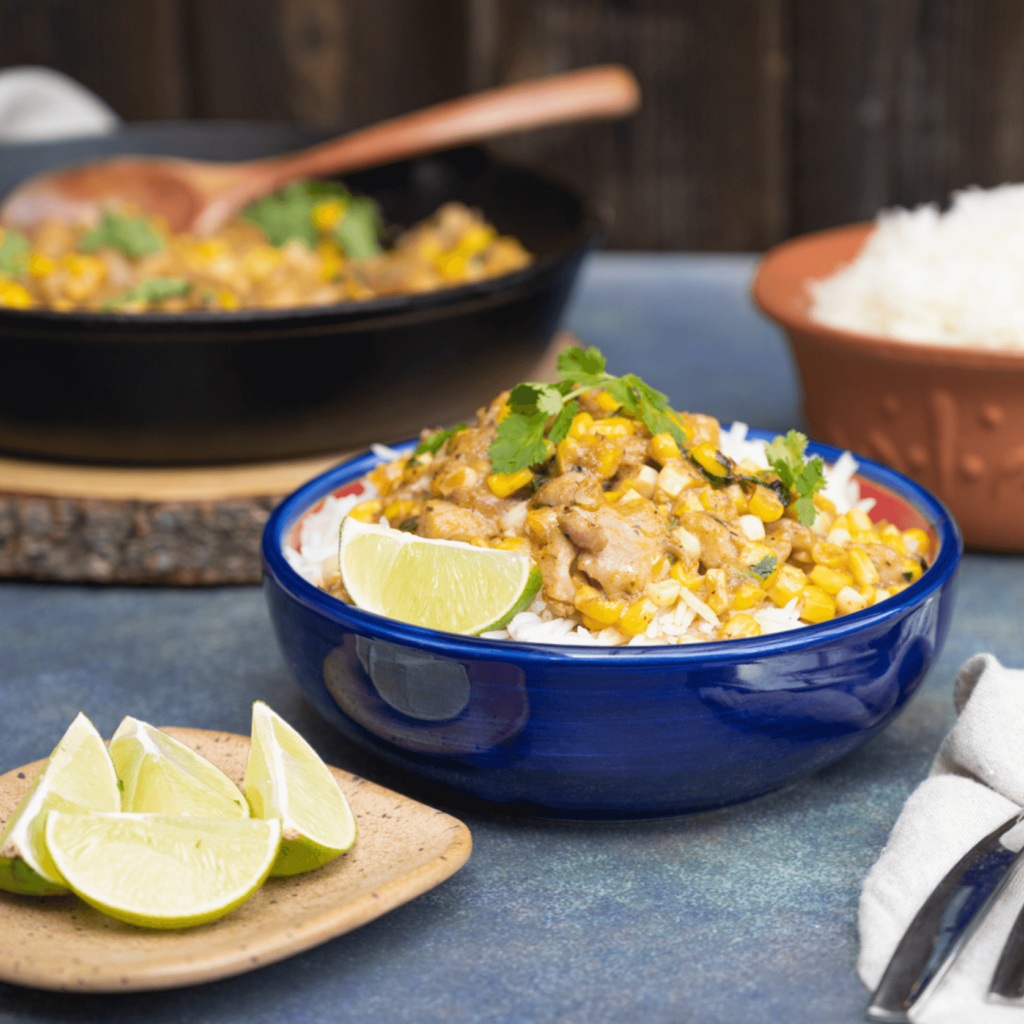 A blue bowl filled with rice and piled high with cooked chicken and fresh corn, garnished with lime wedges and cilantro leaves.