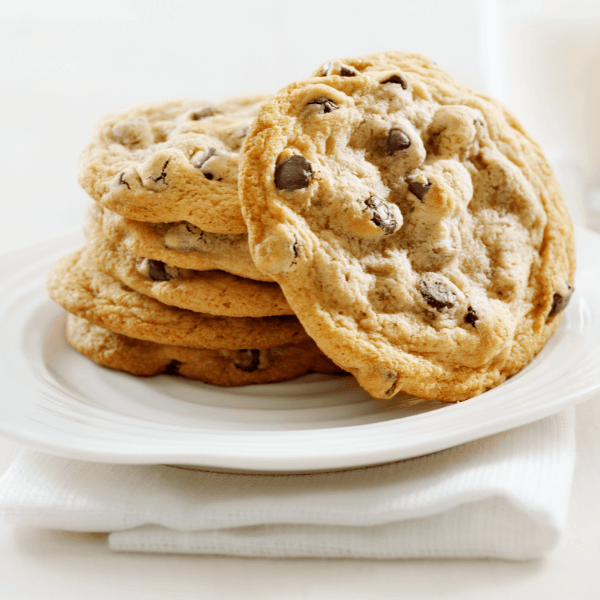 The image shows a white table with a folded white napkin and a white plate on top. On the plate are a stack of five chocolate chip cookies with another cookie angled to face the camera. The cookies are soft and chewy.