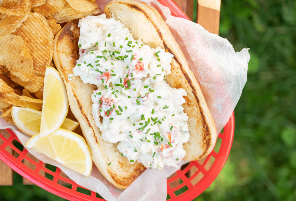 Shot from above, a red plastic basket holds a lobster roll. The bun is toasted and filled with lobster, mayonnaise, and a sprinkle of fresh chives. There are tree wedges of lemon in the basket and golden crinkle cut potato chips.