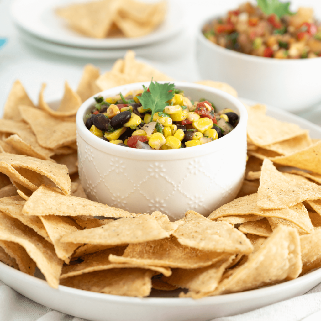 A plate of tortilla chips has a bowl piled high with Farm Boy Roasted Corn and Black Bean Salsa in the centre.