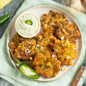 A plate of corn fritters with a small bowl of dipping sauce, garnished with fresh jalapeno peppers.