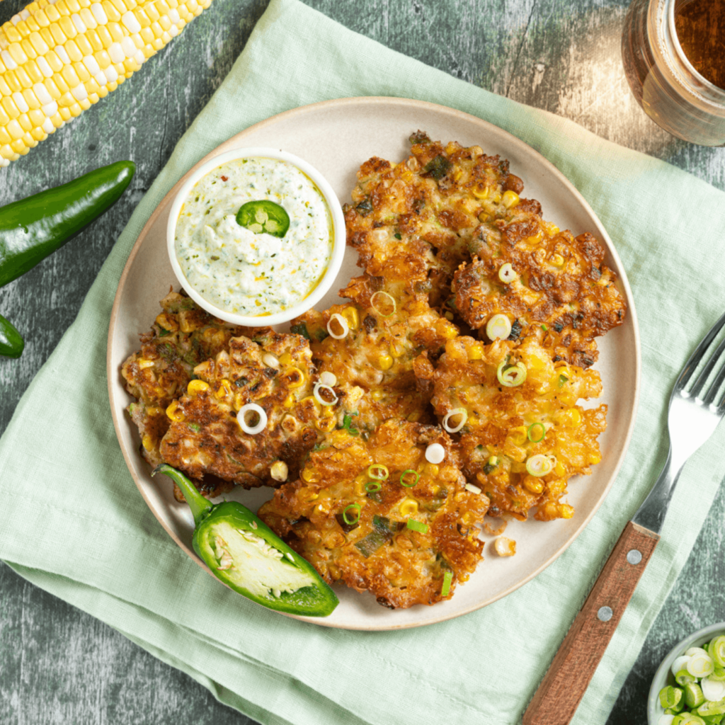 Shot from above, a plate of corn fritters with a small bowl of chimichurri yogurt sauce, garnished with fresh jalapenos.