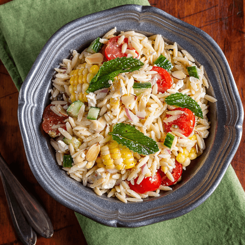 Bowl of fresh orzo salad with corn, cherry tomatoes, almonds, onion, and fresh mint leaves.