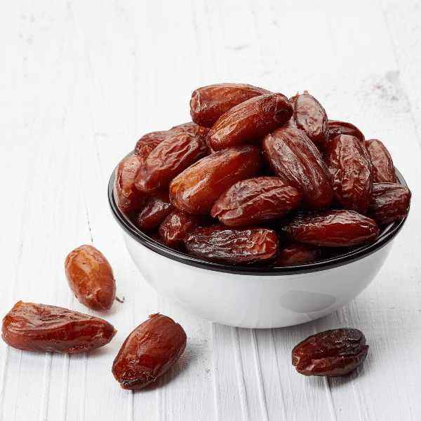 White bowl of dates on white table. Four dates are scattered around the bowl.