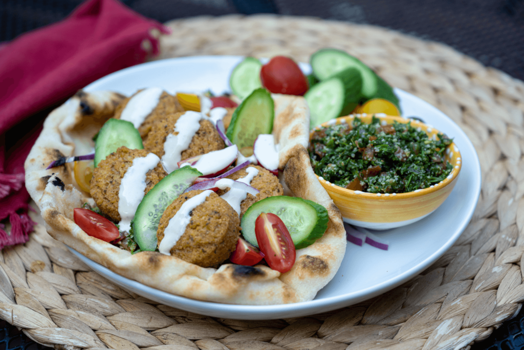 The image shows a white plate on a woven circular placemat. On the plate is a falafel wrap garnished with sliced cucumbers and tahini sauce. There is a small yellow bowl of fresh tabouleh salad beside it.