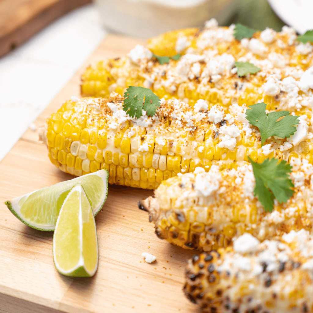 Grilled corn on the cob on a wooden serving tray. Corn is dressed with cotija cheese and cilantro leaves. There are two wedges of lime on the tray.