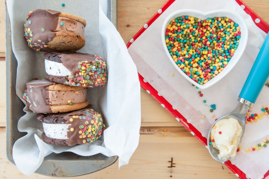 The image shows four ice cream sandwiches dipped in chocolate with rainbow sprinkled lying in a parchment-paper lined loaf pan. To the right side of them is a red and white polka-dot tray with a heart-shaped bowl containing more round rainbow sprinkles and a blue ice cream scoop with vanilla ice cream in it.